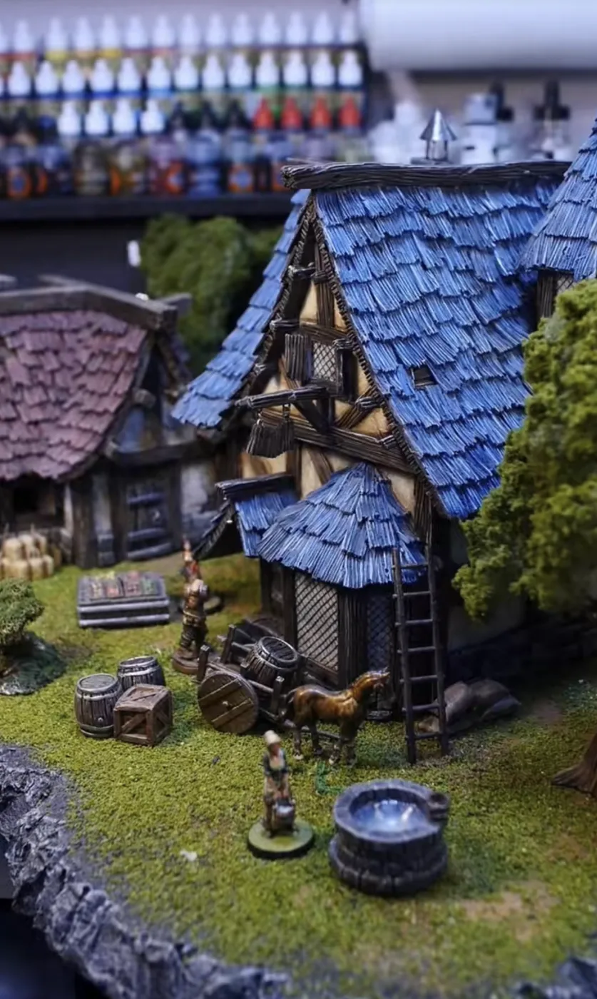 A painted tabletop village: a blue-roofed timber house, barrels, a cart and a well on a grass base.