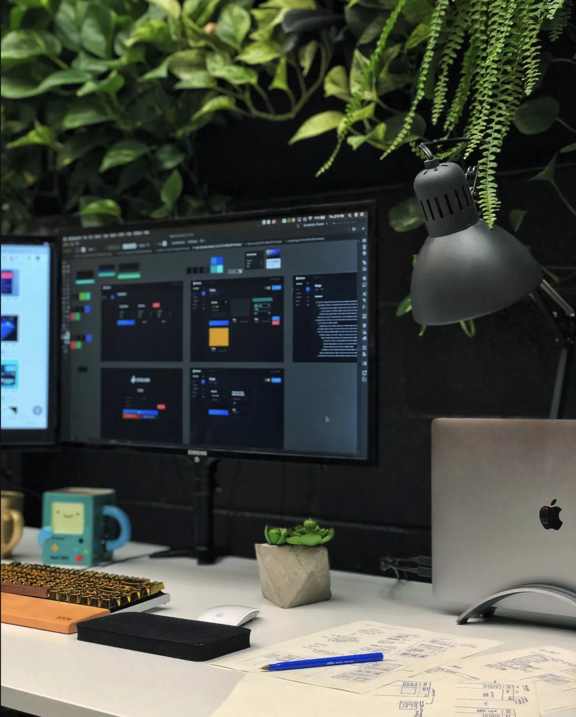 A desk with a monitor showing a dark design system, a laptop, a mechanical keyboard and sketches.